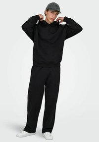 Czarna bawełniana bluza z kapturem i kieszenią kangurką, w połączeniu z czarnymi spodniami dresowymi. Model ma na sobie szarą czapkę i białe trampki.