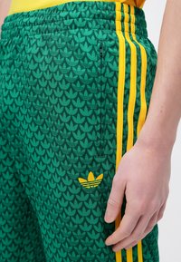 Grün gemusterte Jogginghosen mit gelben Dreistreifen-Akzenten. Verfügt über einen elastischen Bund und ein kleines gelbes Logo am unteren Bein.