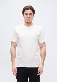 Νέος άνδρας με κοντά σγουρά μαλλιά, που φοράει ένα λευκό T-shirt Columbia και μαύρο παντελόνι, στέκεται μπροστά σε απλό φωτεινό φόντο.