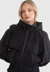 Black hooded jacket with a front zip, featuring a layered design and elastic cuffs. Made of lightweight, smooth material.