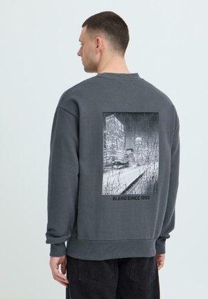 Homme portant un sweat-shirt gris foncé avec une vue de paysage urbain en noir et blanc et le texte "BLEND SINCE 1993" au dos, faisant face à gauche.