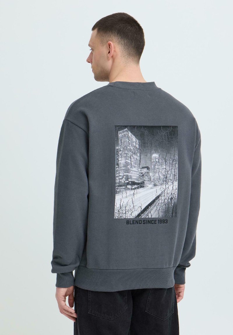 Homme portant un sweat-shirt gris foncé avec une vue de paysage urbain en noir et blanc et le texte "BLEND SINCE 1993" au dos, faisant face à gauche.