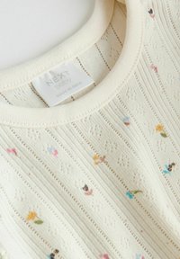 Haut pour bébé couleur crème avec une texture fine en tricot et une broderie florale multicolore dispersée, doté d'une étiquette en tissu au niveau de l'encolure.