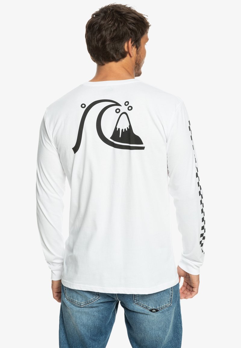 Homme portant une chemise blanche à manches longues avec un graphique de vagues et de montagnes dans le dos et un motif à damier sur la manche droite, associé à un jean bleu.