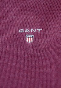 Kaštanová pletená látka s logem "GANT" ve světle šedé barvě a emblémem štítu s červenými a modrými pruhy. Hladká textura s jemným žebrováním.