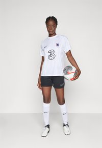 Witte voetbalshirt met reliëfpatroon, zwarte shorts, witte sokken en zwarte voetbalschoenen. Houdt een patroon voetbal vast. Nike-logo zichtbaar.