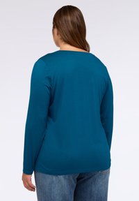 Tílko s dlouhým rukávem v barvě teal s kulatým výstřihem, vyrobené z měkké látky. V kombinaci se světle modrými džíny, zobrazeno ze zadu.