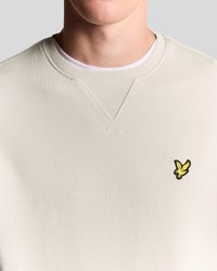 Sudadera beige claro con cuello acanalado, que presenta un pequeño logo bordado en amarillo y negro en la zona izquierda del pecho. Textura suave.