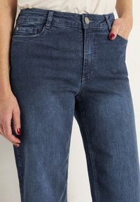 Tummansiniset farkut, joissa on klassinen viiden taskun malli, sileä rakenne ja yksi hopeinen nappi vyötäröllä.