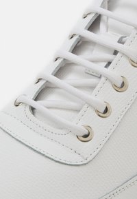 Hvit lær sneakers med strukturert overflate, metalløye, hvite snører og en lys grå aksent langs sålen.