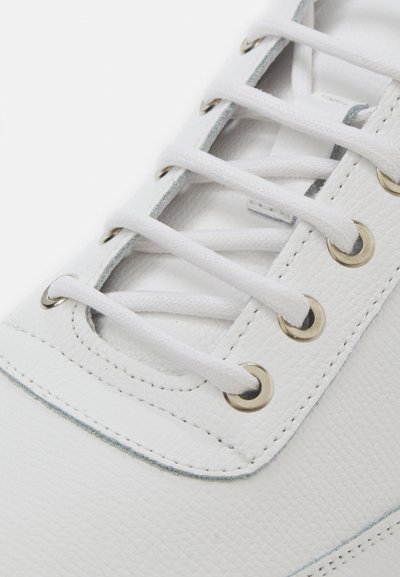 Zapatilla blanca de cuero con superficie texturizada, ojales de metal, cordones blancos y un acento gris claro a lo largo de la suela.