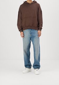 Sudadera con capucha grande de color marrón, con un bolsillo frontal, combinada con unos vaqueros de corte holgado en azul claro y zapatillas blancas. Un conjunto simple y casual.