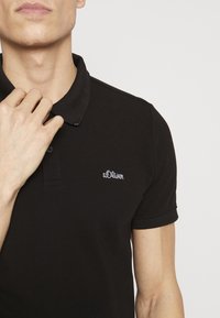 Černé polo tričko vyrobené z texturované tkaniny, s klasickým límečkem a krátkými rukávy. Vyšité logo je umístěno na levé části hrudníku.