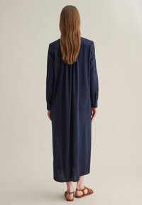 Abito lungo blu navy in tessuto leggero, con un taglio dritto, maniche lunghe e un dettaglio con bottoni sui polsini. Indossato con sandali marroni.