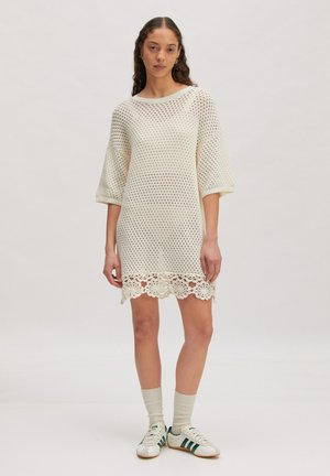 Vrouw staand, gekleed in een crèmekleurige, opengebreide jurk met een bloemenkant zoom, mouwen tot halverwege de arm, witte sokken en witte sneakers met donkere strepen.