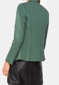 Green fitted blazer with a notch lapel, smooth texture, and tailored silhouette, paired with a black leather skirt.