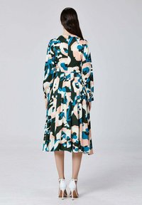 Femme aux longs cheveux bruns portant une robe midi à manches longues à motifs floraux en bleu, rose, blanc et noir, associée à des chaussures à talons blancs, debout.