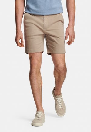 Man wearing beige shorts, beige sneakers, and a light blue shirt, walking with one foot slightly raised against a white background.