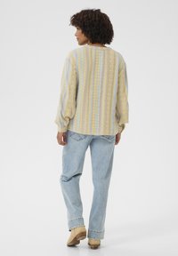 Blouse légère à manches longues de couleur jaune pastel et bleue, ornée de motifs géométriques. Associée à un jean bleu clair à coupe décontractée et des chaussures beiges.