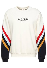 Felpa crema con polsini e fondo a coste neri. Presenta audaci strisce diagonali in rosso, blu, bianco, giallo e nero. Il testo sul davanti recita "KAOTIKO SINCE 1999."