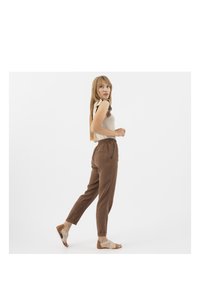 Hnědé, lehké kalhoty s elastickým pasem, zúženými nohavicemi, bočními kapsami a měkkou texturou. Kombinované s sandály s tkaným designem.