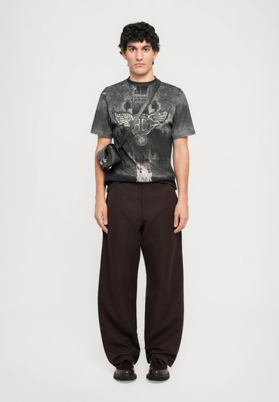 Young man wearing a dark washed graphic t-shirt, loose dark brown pants, black crossbody bag, and dark polished shoes, standing against white background.