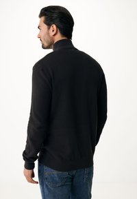 Pullover nero a maglia texturizzata con colletto alto e polsini a coste, abbinato a jeans in denim blu, visto da dietro.