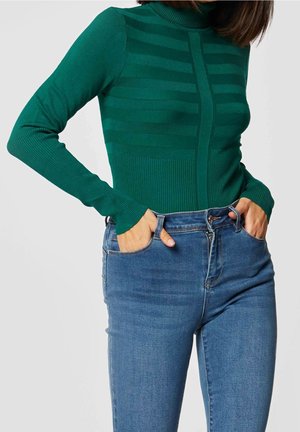 Frau trägt einen eng anliegenden dunkelgrünen gestreiften Pullover und hoch taillierte blaue Jeans, die Hände in den Taschen versteckt.