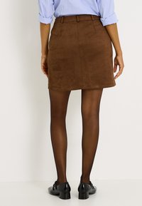 Minifalda marrón de ante sintético con corte recto y dos bolsillos traseros, combinada con medias negras transparentes y calzado negro brillante.