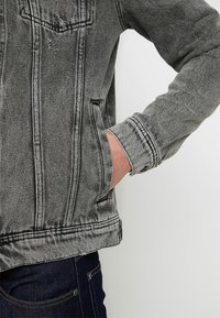 Šedá džínová bunda s opotřebovaným vzhledem, klasickým límcem, předními kapsami a střih na tělo. Má kontrastní švy a žebrované manžety.