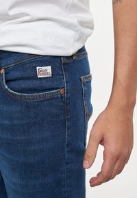 Jeans in denim blu con un design aderente, dotati di un'etichetta bianca e rossa sulla tasca posteriore e cuciture arancioni a contrasto.