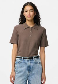 Brun pikétröja med knappslå, korta ärmar och en slät textur, i kombination med ljusblå högmidjade jeans och ett svart bälte.