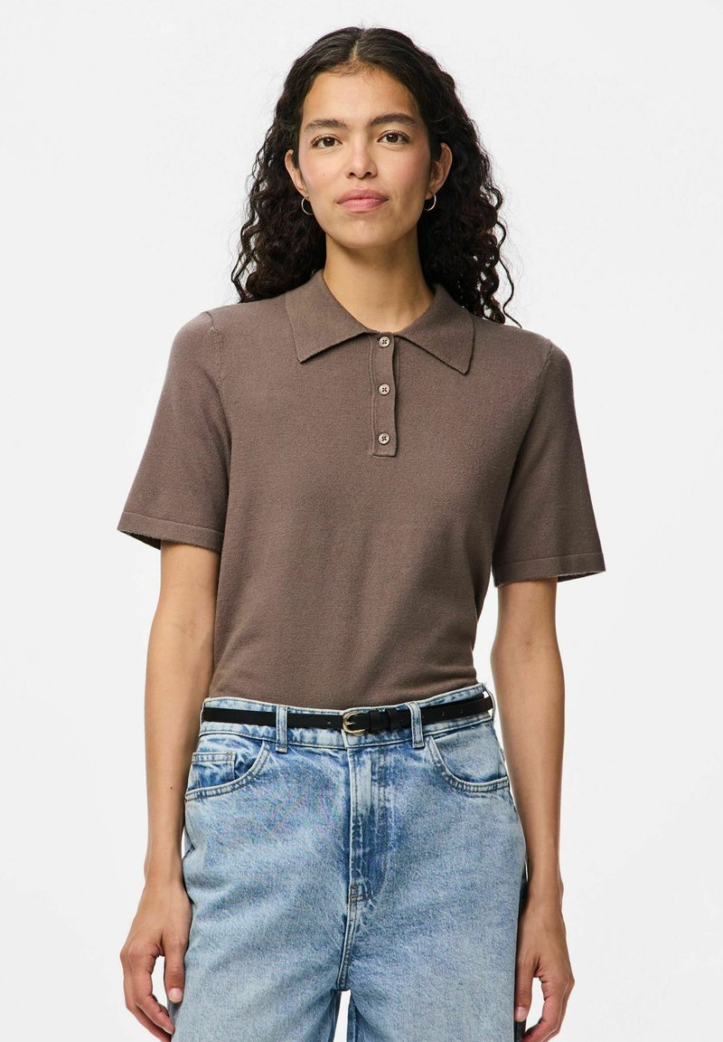 Brun pikétröja med knappslå, korta ärmar och en slät textur, i kombination med ljusblå högmidjade jeans och ett svart bälte.