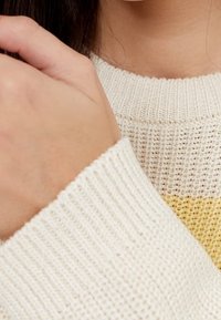 Close-up van een persoon die een crème kleurige gebreide trui met een gele streep draagt, hand rustend nabij de nek en donker haar zichtbaar.