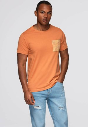 Kortærmet orange t-shirt med en struktureret lysebrun lomme, parret med lyseblå slidte jeans med en afslappet pasform.