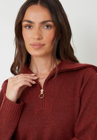 Vrouw met lang bruin haar, draagt een rode gebreide trui met een gouden rits en een gouden ketting, die de ketting voorzichtig aanraakt.