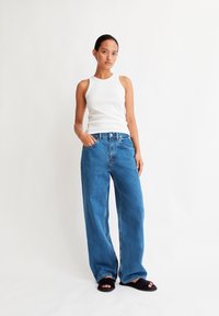 A woman stands confidently in a sleeveless white top and wide-leg blue jeans, hands on her hips, against a plain white background.