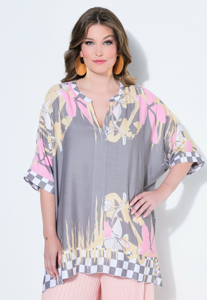 Vrouw met golvend bruin haar draagt een oversized grijze tuniek met roze, beige bloemenprint en geruite rand, gecombineerd met roze geplooide broek.