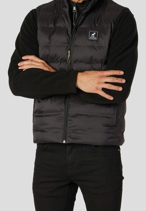 Mężczyzna noszący czarną kamizelkę puchową z przednim zamkiem błyskawicznym i naszywką z logo, z założonymi na piersi rękami na czarnej koszulce z długim rękawem i czarnych spodniach.