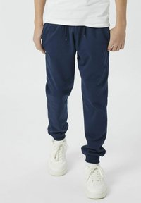 Pantalons de jogging bleu marine avec des poignets élastiqués, une taille à cordon de serrage et des poches latérales, fabriqués dans un tissu doux et léger. Associés à des baskets blanches.