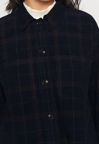 Giacca camicia in corduroy navy con motivo a quadri in rosso scuro, colletto a punta e chiusura centrale con bottoni. Due tasche frontali visibili.