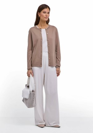 Donna in piedi, indossa un cardigan doppiopetto color taupe sopra una maglietta bianca e pantaloni bianchi ampi, con una borsa a spalla a quadri e una sciarpa bianca.