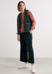 Žalia flisinė liemenė su dviem kišenėmis, dėvima virš raudonos ir baltos horizontaliai dryžuotos ilgomis rankovėmis marškinėlių. Derinama su tamsiai žaliais aksominiais plačiais keliais ir baltais sportbačiais.