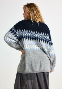 Donna con capelli biondi ricci che indossa un maglione largo grigio, blu navy e blu con un motivo, in piedi di lato con la mano sulla vita contro uno sfondo bianco.