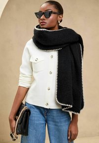 Fular texturat negru cu margine albă în zigzag, asortat cu un pulover alb cu nasturi și blugi. Accesoriile includ o geantă neagră și ochelari de soare.