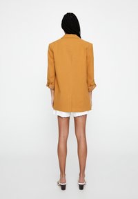 Blazer jaune moutarde avec une coupe décontractée, manches retroussables et une fente au dos. Associé à un short blanc déchiré et des sandales à talons carrés.