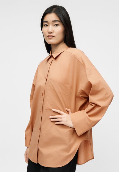 Mladá žena s dlhými tmavými vlasmi, ktorá má na sebe nadrozmernú svetlohnedú košeľu na gombíky s predným vreckom a čierne nohavice.