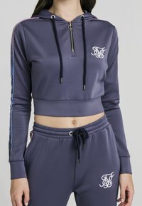 Fialová krátká mikina s kapucí na zip, tmavě modrým šňůrkem a logem na hrudi; doplněná o sladěné tepláky s podobnými designovými prvky.