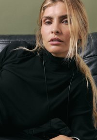 Jersey negro de cuello alto hecho de una tela suave, con un diseño ajustado. Se aprecian largos cabellos rubios ondulados y accesorios mínimos.