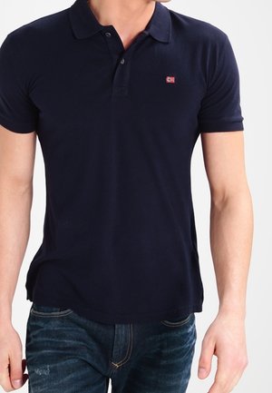 Mann trägt ein marineblaues Poloshirt mit kurzen Ärmeln, einem kleinen rot-weißem Flaggenlogo auf der linken Brust und dunkelblaue Jeans.